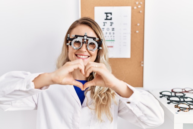 Optician woman wearing optometry glasses at the clinic smiling showing heart shape with hands.